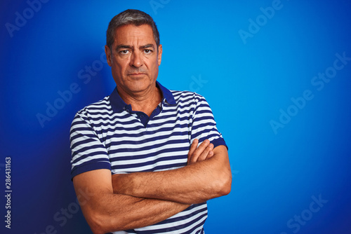Handsome middle age man wearing striped polo standing over isolated blue background skeptic and nervous, disapproving expression on face with crossed arms. Negative person.