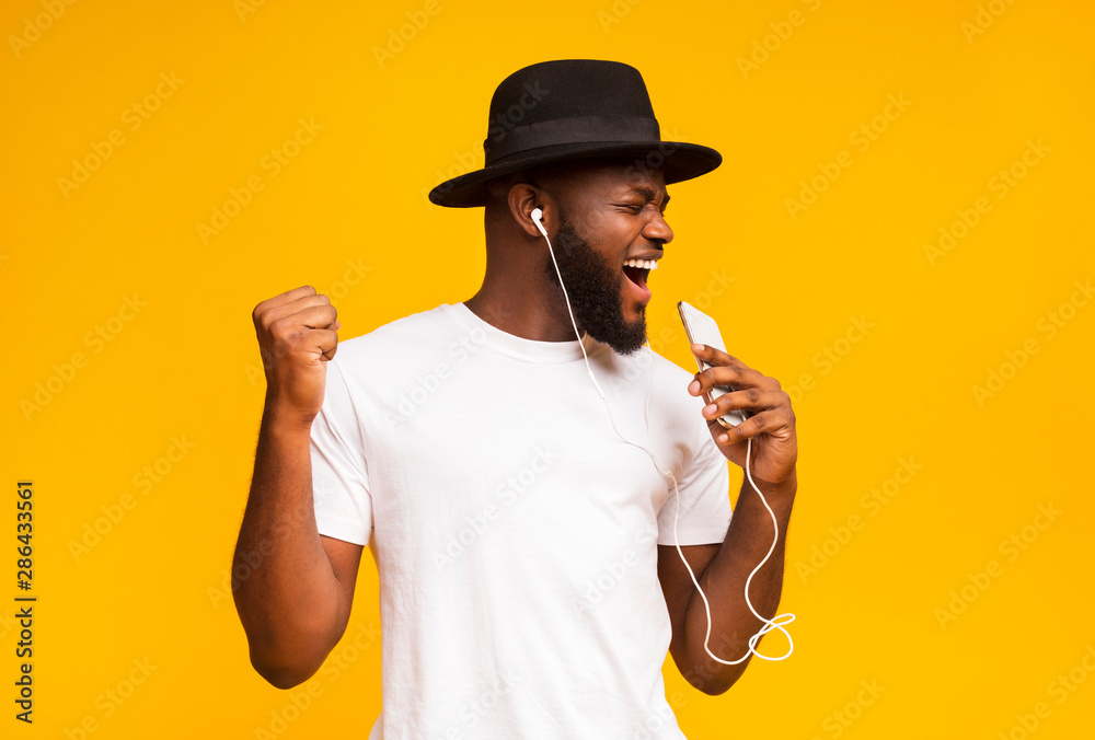 © Prostock-studio - Happy african man in hat singing into smartphone like microphone