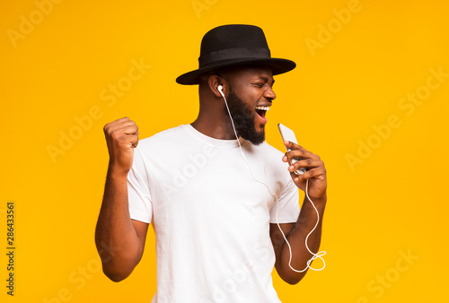 Obraz na plátně Happy african man in hat singing into smartphone like microphone