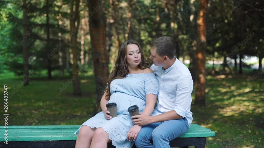 Pregnant Woman and her Husband Drinking Tea and Talking while Sitting on the Bench in the Park. Slow Motion. The Concept of Family Happiness Pregnant Woman and her Husband Drinking Tea and Talking while Sitting on the Bench in the Park. Slow Motion. The Concept of Family Happiness
