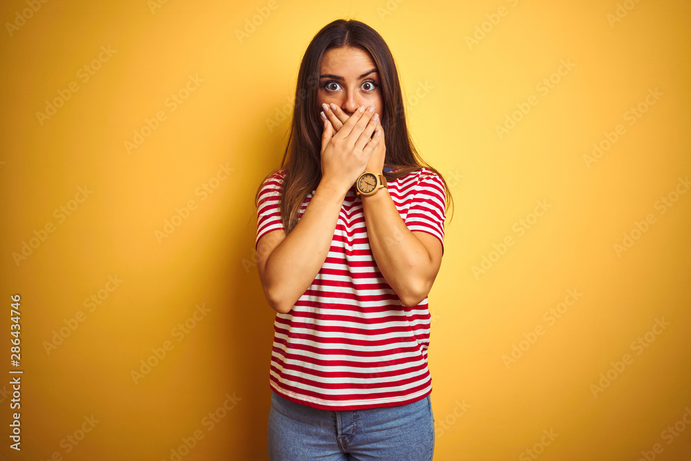 Young beautiful woman wearing striped t-shirt standing over isolated yellow background shocked covering mouth with hands for mistake. Secret concept. Young beautiful woman wearing striped t-shirt standing over isolated yellow background shocked covering mouth with hands for mistake. Secret concept.