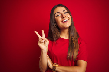 Young beautiful woman wearing t-shirt standing over isolated red background smiling with happy face winking at the camera doing victory sign. Number two.
