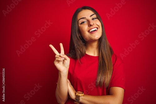 Young beautiful woman wearing t-shirt standing over isolated red background smiling with happy face winking at the camera doing victory sign. Number two. photo