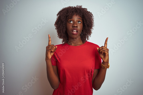Young african afro woman wearing red t-shirt over isolated white background amazed and surprised looking up and pointing with fingers and raised arms.
