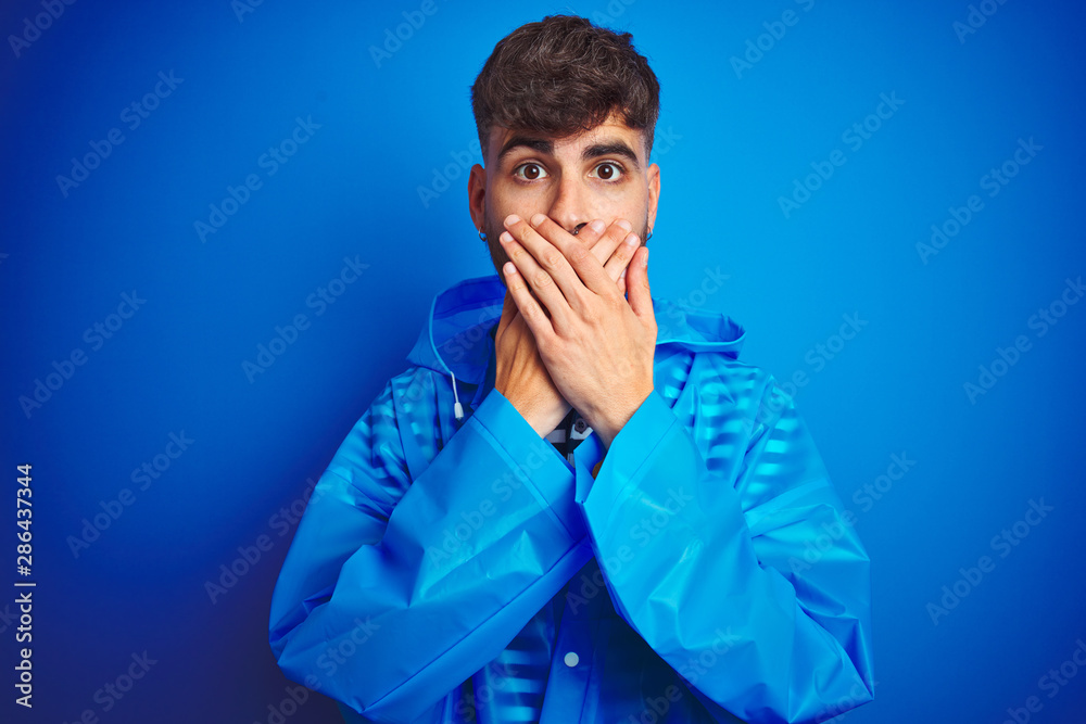 Young handsome man wearing rain coat standing over isolated blue background shocked covering mouth with hands for mistake. Secret concept. Young handsome man wearing rain coat standing over isolated blue background shocked covering mouth with hands for mistake. Secret concept.