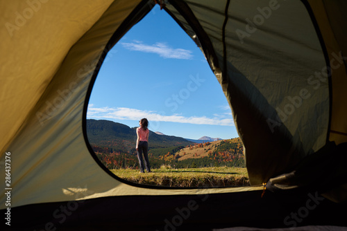 View from inside of hikers tourist tent in mountains. Slim sportive dark-haired young woman standing on grassy valley on background of distant hills and bright blue sky on sunny summer day.