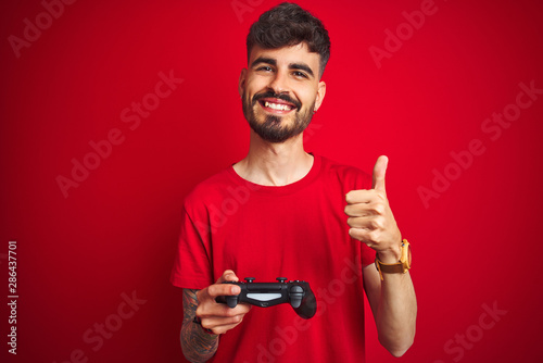 Young gamer man with tattoo playing video game standing over isolated red background happy with big smile doing ok sign, thumb up with fingers, excellent sign