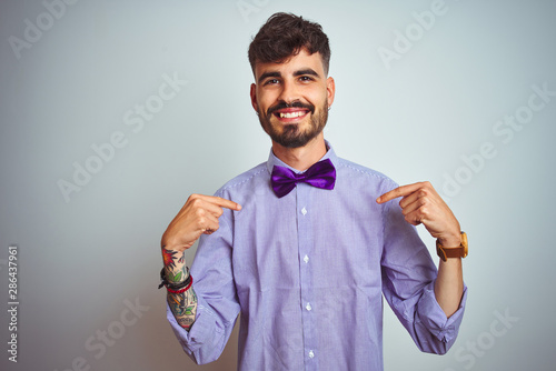 Young man with tattoo wearing purple shirt and bow tie over isolated white background looking confident with smile on face, pointing oneself with fingers proud and happy.