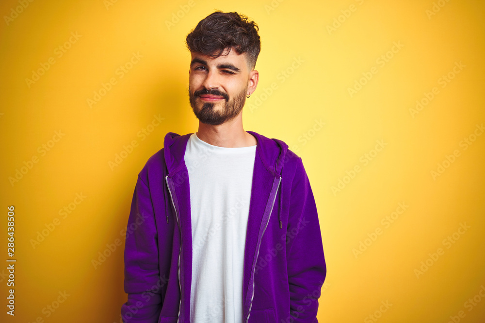 Young man with tattoo wearing sport purple sweatshirt over isolated yellow background winking looking at the camera with sexy expression, cheerful and happy face.