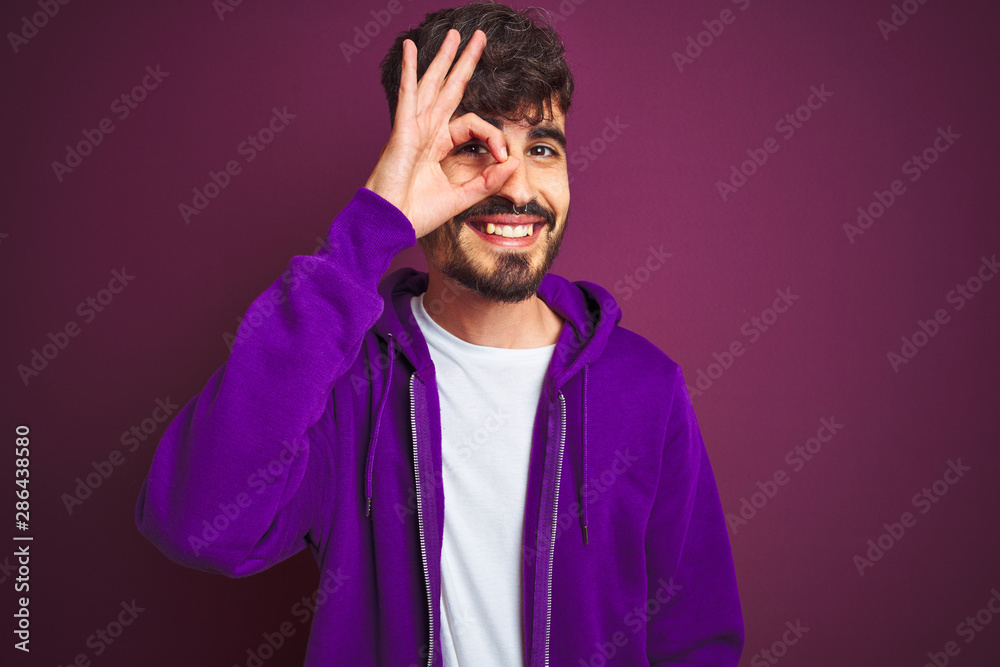 Young man with tattoo wearing striped polo standing over isolated yellow background doing ok gesture with hand smiling, eye looking through fingers with happy face. Young man with tattoo wearing striped polo standing over isolated yellow background doing ok gesture with hand smiling, eye looking through fingers with happy face.