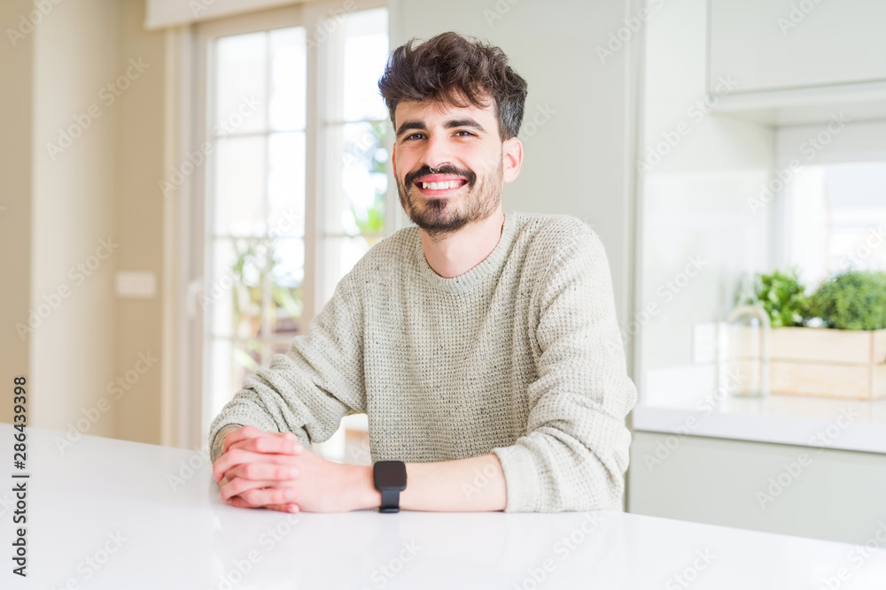 Young man wearing casual sweater sitting on white table with a happy and cool smile on face. Lucky person.