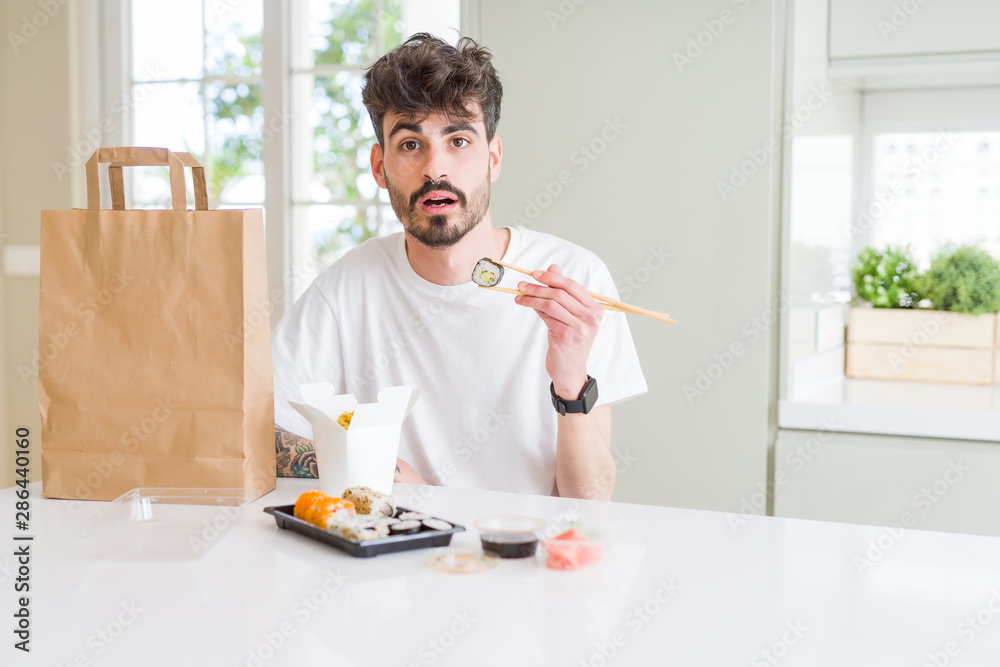 Young man eating asian sushi from home delivery scared in shock with a surprise face, afraid and excited with fear expression Young man eating asian sushi from home delivery scared in shock with a surprise face, afraid and excited with fear expression