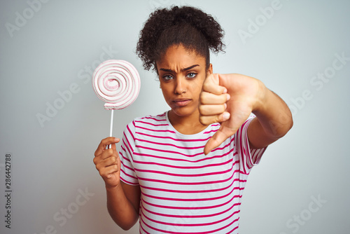 African american teenager woman eating colorful candy over isolated white background with angry face, negative sign showing dislike with thumbs down, rejection concept