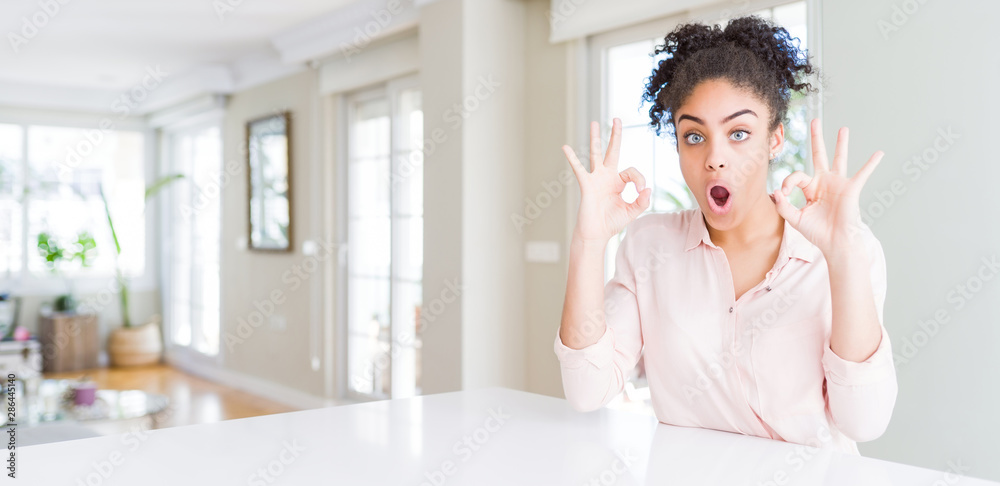 Wide angle of beautiful african american woman with afro hair looking surprised and shocked doing ok approval symbol with fingers. Crazy expression