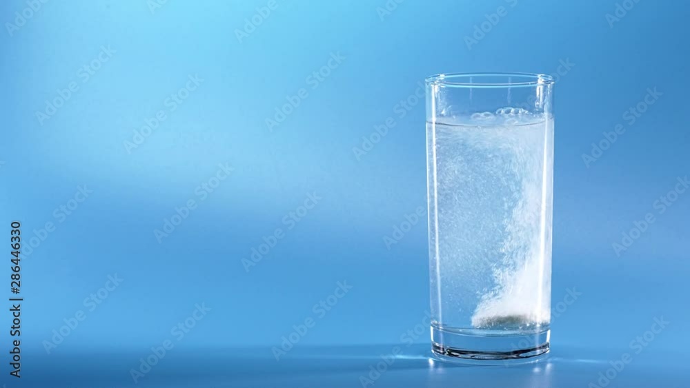 Effervescent Tablet Falling In Water. Pill Falls And Dissolves With Bubbles. Effervescent Tablet In Glass Of Water On Blue Background. Shot Of Effervescent Pill Dissolving In A Glass Of Water. Effervescent Tablet Falling In Water. Pill Falls And Dissolves With Bubbles. Effervescent Tablet In Glass Of Water On Blue Background. Shot Of Effervescent Pill Dissolving In A Glass Of Water.