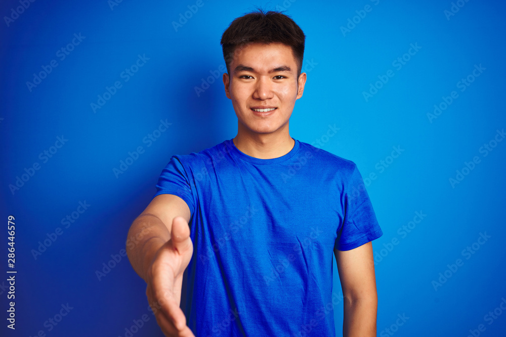 Young asian chinese man wearing t-shirt standing over isolated blue background smiling friendly offering handshake as greeting and welcoming. Successful business. Young asian chinese man wearing t-shirt standing over isolated blue background smiling friendly offering handshake as greeting and welcoming. Successful business.