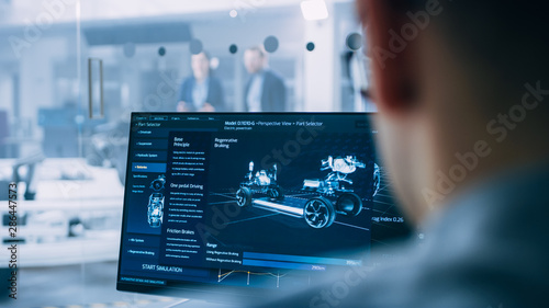Professional Engineer Works on a Computer with a 3D CAD Software and Tests the Electric Car Chassis Prototype with Wheels, Batteries and Engine Standing in a High Tech Development Laboratory. photo