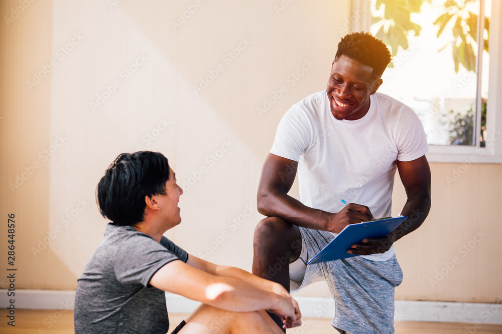 Obraz premium African American fitness instructor with clipboard writing down results of exhausted sportsman sitting on floor exercising and smiling