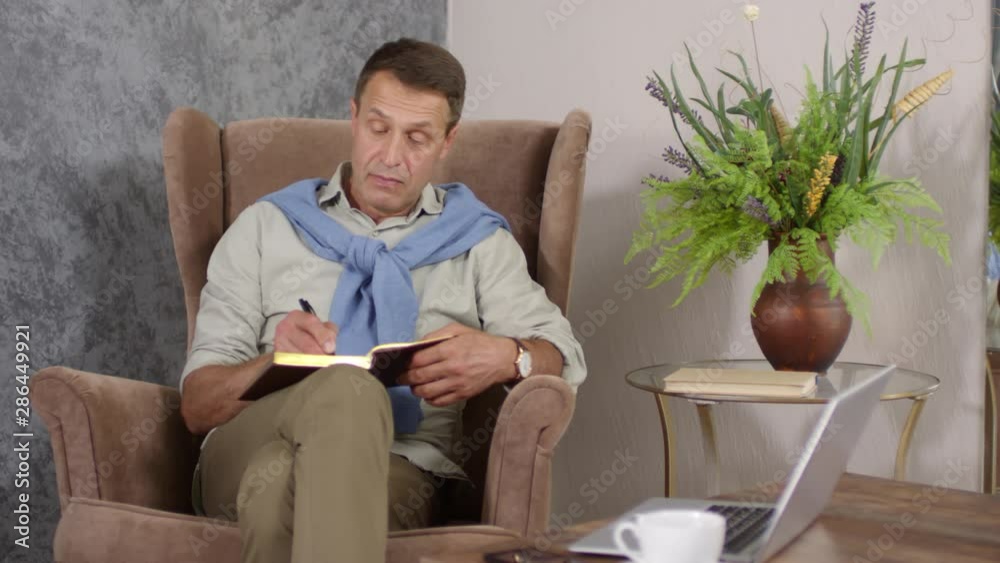 Knee-up shot of middle-aged good-looking thoughtful Caucasian man sitting in armchair at home and writing in journal, with bouquet of flowers, laptop and coffee on table nearby Knee-up shot of middle-aged good-looking thoughtful Caucasian man sitting in armchair at home and writing in journal, with bouquet of flowers, laptop and coffee on table nearby