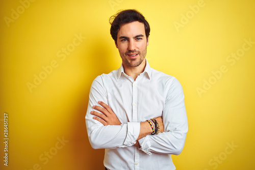Young handsome businessman wearing elegant shirt standing over isolated yellow background happy face smiling with crossed arms looking at the camera. Positive person.