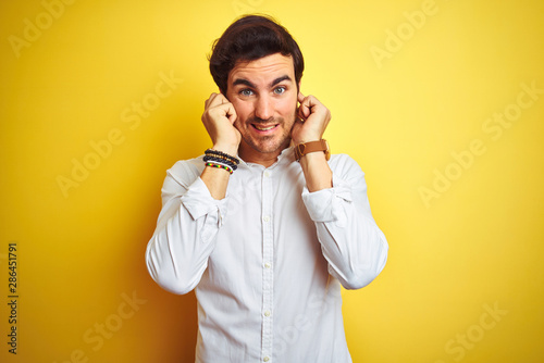 Young handsome businessman wearing elegant shirt standing over isolated yellow background covering ears with fingers with annoyed expression for the noise of loud music. Deaf concept.