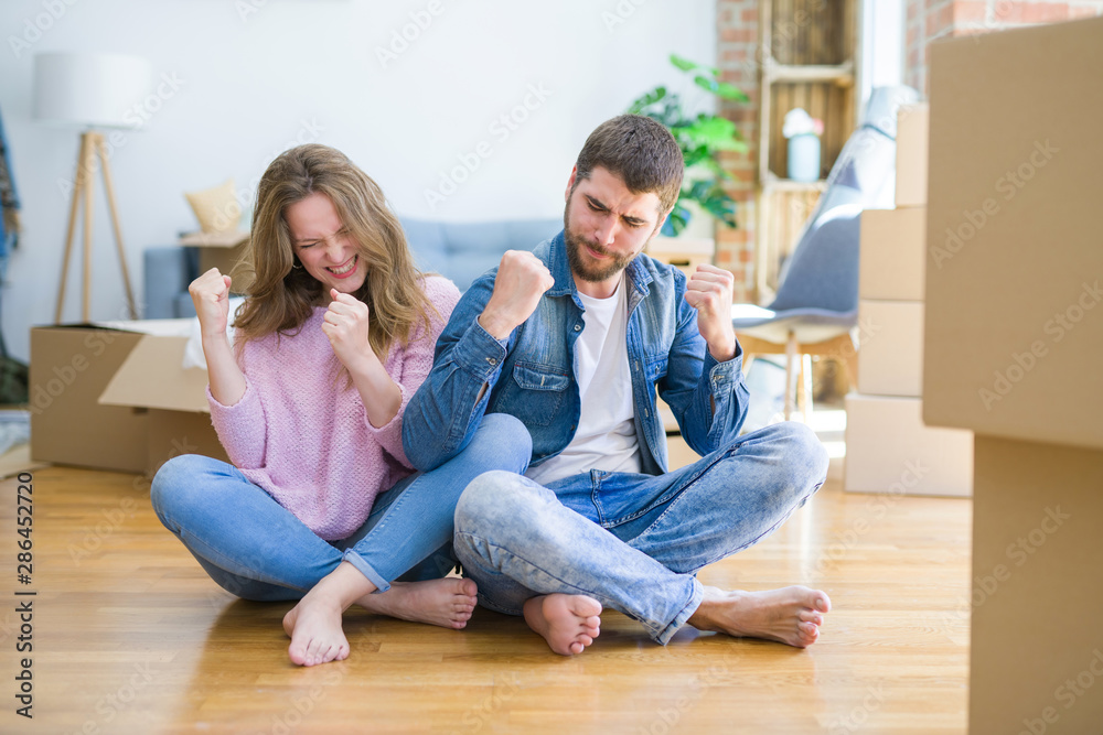 Young beautiful couple moving to a new house sitting on the floor very happy and excited doing winner gesture with arms raised, smiling and screaming for success. Celebration concept. Young beautiful couple moving to a new house sitting on the floor very happy and excited doing winner gesture with arms raised, smiling and screaming for success. Celebration concept.