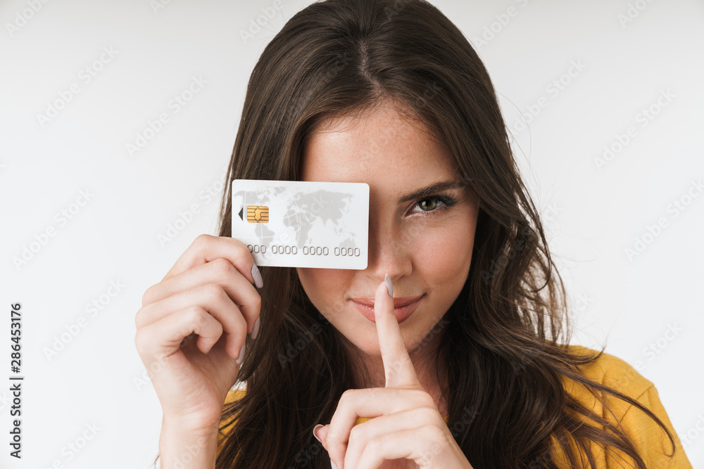 Image of beautiful brunette woman wearing casual clothes keeping finger at lips and holding credit card - obrazy, fototapety, plakaty 
