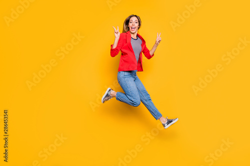 Full body photo of lovely girl jumping making v-sign wearing red striped shirt jacket denim jeans isolated over yellow background
