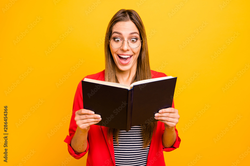 Portrait of astonished hipster with long hair holding printed fiction book wearing eyeglasses eyewear isolated over yellow background