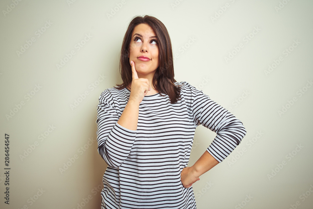 Young beautiful brunette woman wearing striped sweater over white isolated background with hand on chin thinking about question, pensive expression. Smiling with thoughtful face. Doubt concept. Young beautiful brunette woman wearing striped sweater over white isolated background with hand on chin thinking about question, pensive expression. Smiling with thoughtful face. Doubt concept.