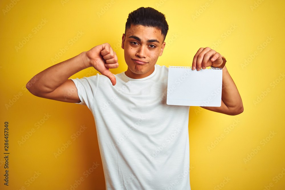 Young brazilian man showing paper banner standing over isolated yellow background with angry face, negative sign showing dislike with thumbs down, rejection concept Young brazilian man showing paper banner standing over isolated yellow background with angry face, negative sign showing dislike with thumbs down, rejection concept