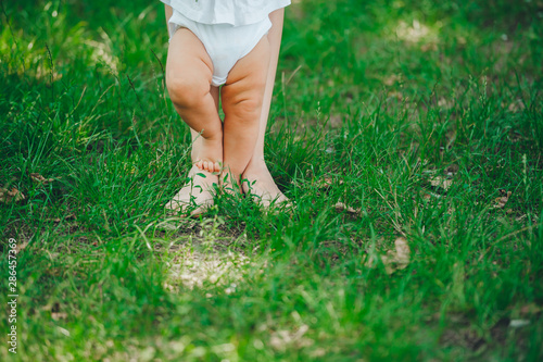 First steps. Mom and daughter's feet on green grass in summer park.