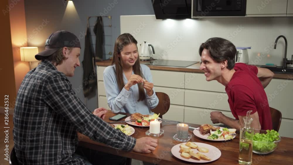 Three cheerful young friends, two men and woman, sitting at table in kitchen, having dinner and talking in evening Three cheerful young friends, two men and woman, sitting at table in kitchen, having dinner and talking in evening