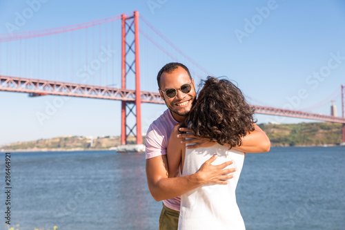 Joyful handsome Latin man embracing girlfriend outdoors. Happy couple standing near river and hugging. Romantic walk concept