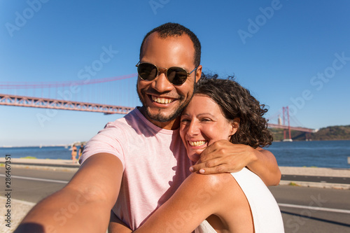 Couple of joyful tourists enjoying vacation and taking selfie on city promenade. Happy man and woman standing outside, holding smartphone and hugging. Happy couple concept © Mangostar