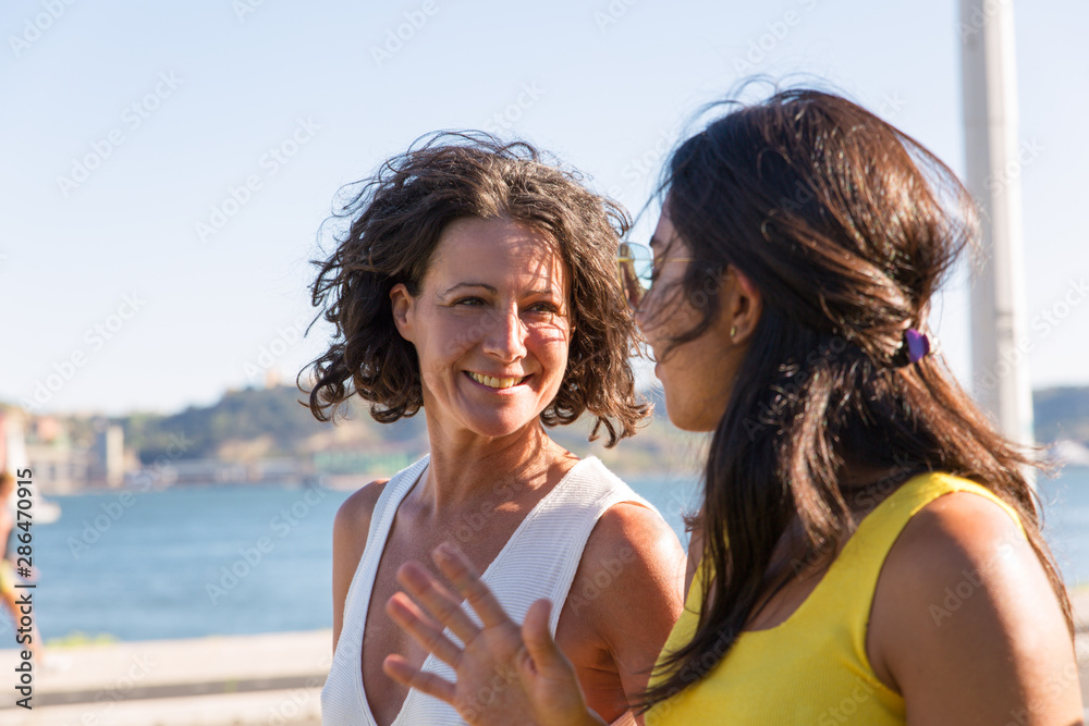 Happy female friends walking on promenade and chatting. Two women meeting outdoors, talking and laughing. Female friendship concept Happy female friends walking on promenade and chatting. Two women meeting outdoors, talking and laughing. Female friendship concept