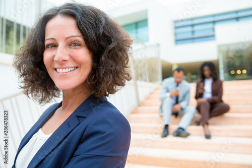 Cheerful middle aged businesswoman. Portrait of happy businesswoman smiling at camera while coworkers sitting on steps behind. Business concept