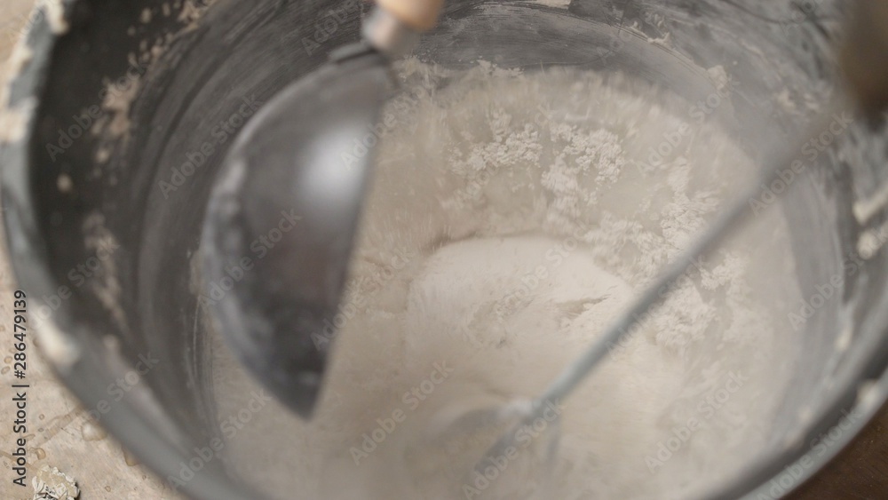 Mix the solution in a bucket using a construction mixer. Worker mixing plaster in a bucket for alignment and putty walls of the apartment using an electric drill, close-up. Mix the solution in a bucket using a construction mixer. Worker mixing plaster in a bucket for alignment and putty walls of the apartment using an electric drill, close-up.