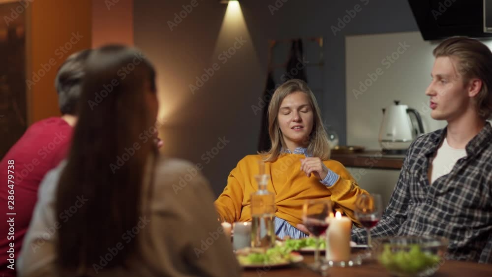 Group of four young friends, two men and two women, sitting at table in domestic kitchen and talking over dinner and wine. Young man speaking. Focus on woman listening attentively to boyfriend Group of four young friends, two men and two women, sitting at table in domestic kitchen and talking over dinner and wine. Young man speaking. Focus on woman listening attentively to boyfriend