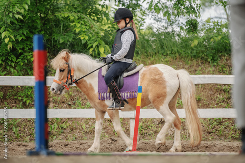 Caucasian girl with helmet and protective vest on riding cute white and brown pony horse. Sunny day on ranch concept.