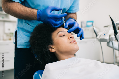 Attractive young African woman is getting a rejuvenating facial injections. She is sitting calmly at clinic. The expert beautician is filling female wrinkles by hyaluronic acid.