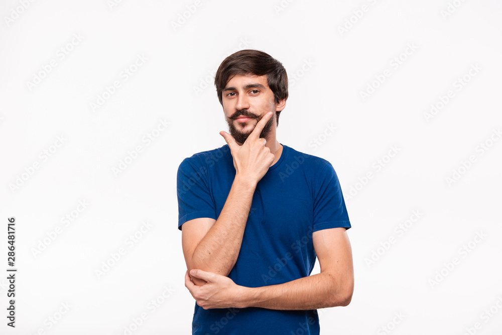 Thoughtful handsome brunet man in a blue shirt holding arms folded over his chest and keeping one hand close to face standing isolated over white background. Thoughtful handsome brunet man in a blue shirt holding arms folded over his chest and keeping one hand close to face standing isolated over white background.