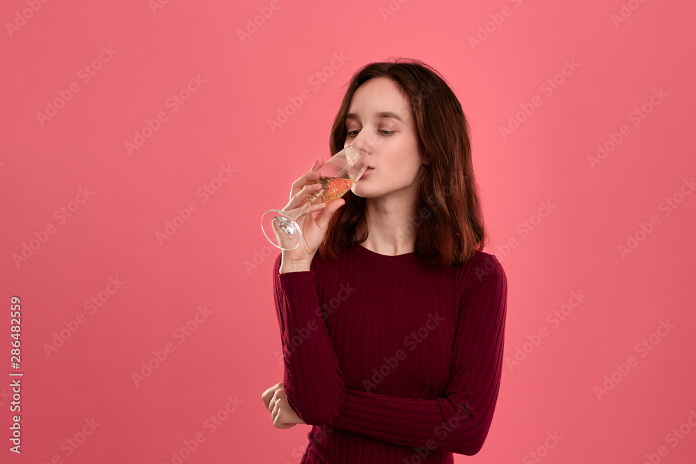 Excited pretty brunette girl drinking sparkling champagne from a wineglass standing isolated on a dark pink background. Excited pretty brunette girl drinking sparkling champagne from a wineglass standing isolated on a dark pink background.