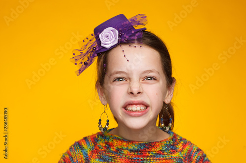 Portrait of a cheerful 8-year-old girl in a miniature hat and in a multi-colored sweater. Yellow background