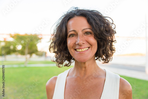 Happy female tourist enjoying outdoor walk in park. Middle aged woman in casual standing on grass and smiling at camera. Outdoor female portrait concept