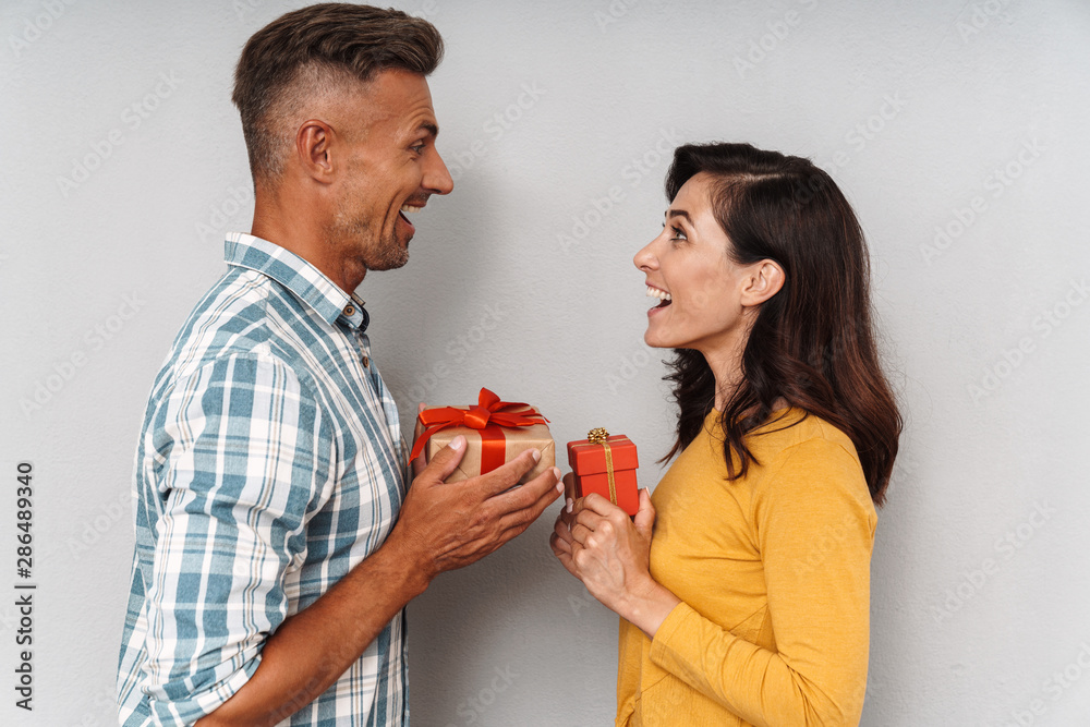 Cute optimistic adult loving couple isolated over grey wall background holding gifts for each other. Cute optimistic adult loving couple isolated over grey wall background holding gifts for each other.
