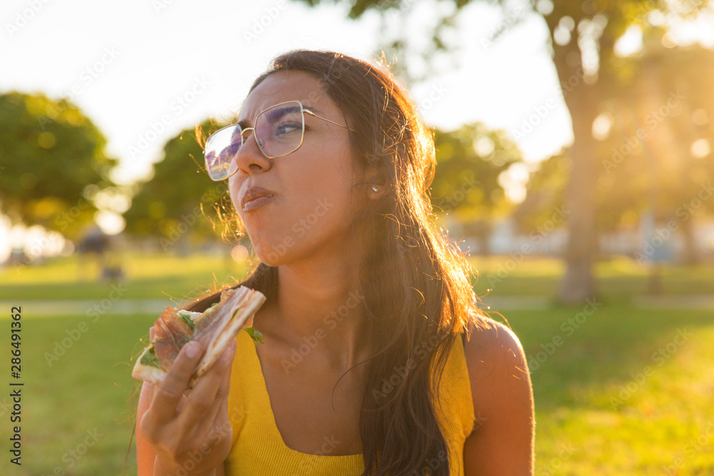 Happy delighted Latin woman enjoying tasty pizza outdoors. Beautiful young woman sitting on grass in park and eating pizza. Tasty food concept