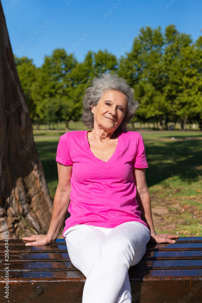 Peaceful old lady posing in park. Senior grey haired woman in casual sitting on bench, leaning on hands and looking at camera. Old woman in park concept