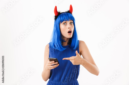 Portrait of cute perplexed woman wearing blue wig and toy devil horns pointing finger at her cellphone