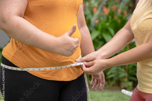 Chuby woman raised her hand to show the satisfaction sign that she had successfully lost weight.  Her friend used a tape measure to the waist size in the front garden at home. Concept about self-care. photo